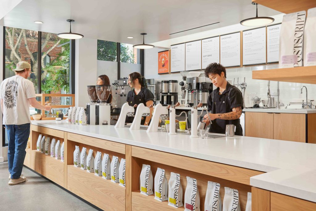Postscript coffee bar interior with baristas brewing espresso behind a white oak service counter, San Francisco, CA