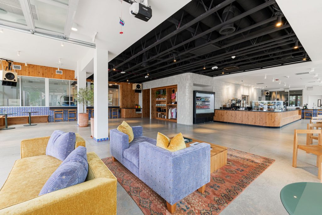 Interior of Desert Kid Coffee, Palm Desert, CA. A blue patterned sofa and mustard armchair sit on a Persian rug in the foreground. Behind them, the full café stretches back — retail shelving, a refrigerated grab-and-go case, the main bar, and the slow bar. Exposed black steel ceiling trusses run the length of the space. Natural light from tall windows on the left. Polished concrete floors throughout.